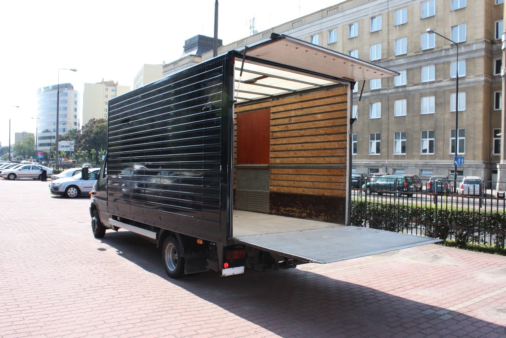 Czarny samochód dostawczy z otwartą windą załadunkową na tle miejskiego budynku z parkującymi samochodami.